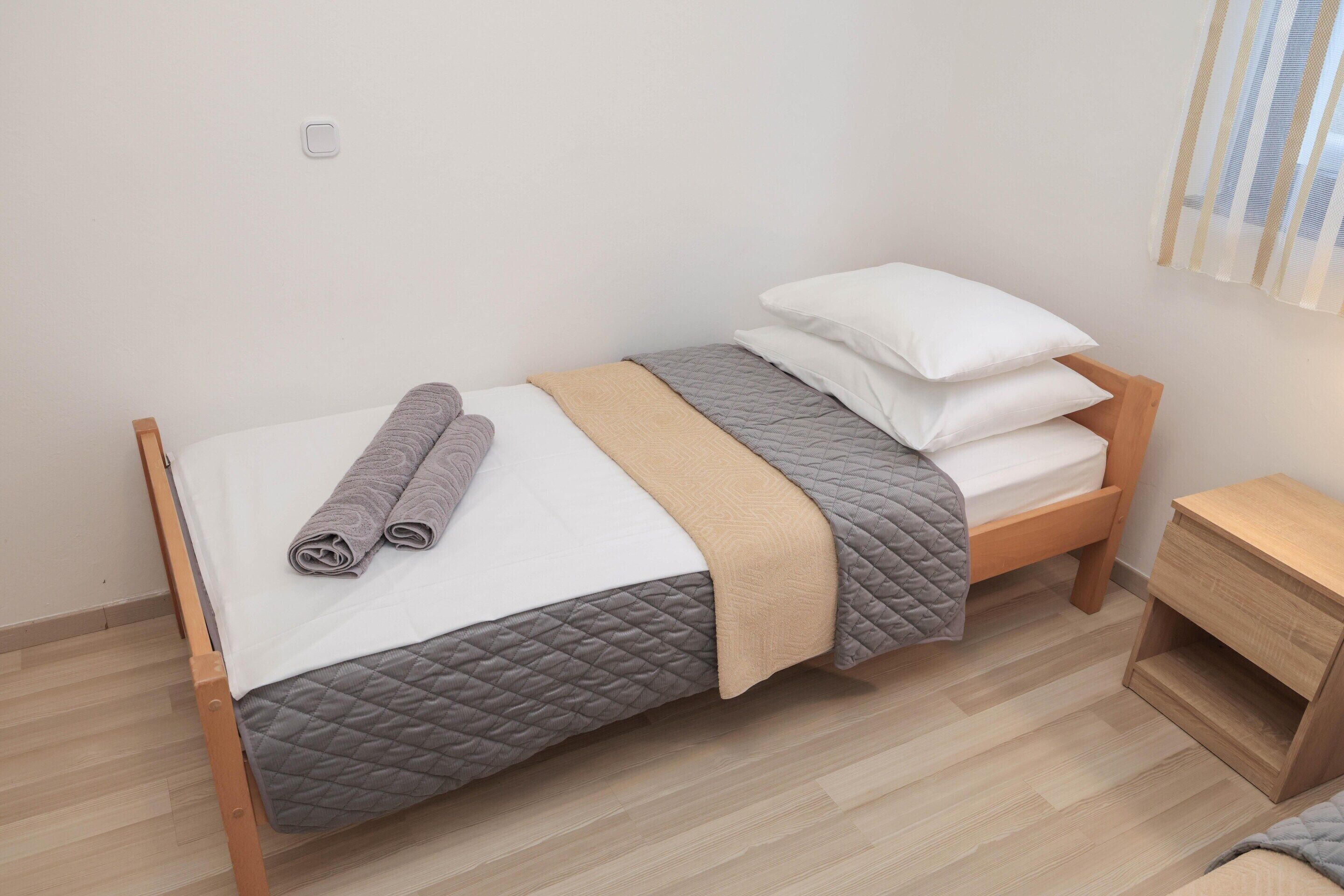 Ein Einzelbett mit gesteppter Bettdecke, zusammengerollte Handtücher und ein Nachttisch mit Holzoptik.