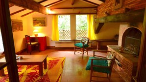 A cozy living room with a fireplace, wooden beams, a table, chairs, and a view through a window with curtains. - Peaceful Retreat With Shared Pool (San Marcello Piteglio)