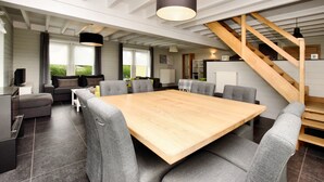 A spacious room with a large wooden table surrounded by grey upholstered chairs, a staircase, and a kitchen area in the background. - Holiday Home Suitable for Families With Sauna (Houffalize)