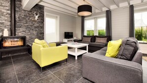 A modern living room with a stone fireplace, a sofa, a yellow armchair, and a television. - Holiday Home Suitable for Families With Sauna (Houffalize)