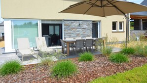 A modern house with a patio area featuring a table and chairs, surrounded by a garden with various plants and a large umbrella. - Holiday Home Suitable for Families With Sauna (Houffalize)
