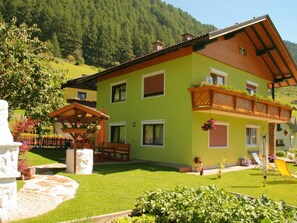 A two-story house with a green facade, wooden balcony, and a stone pathway leading to a wooden pergola. - Newly Furnished Appartment at the Mouth of the Poller Valley National Park (Rennweg am Katschberg)