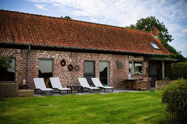 Un bâtiment en briques surmonté d’un toit en tuiles, avec deux fauteuils de jardin, une petite table et un espace aménagé en jardin.
