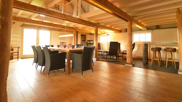 Quarto espaçoso com teto alto, vigas de madeira, área de jantar com cadeiras e uma mesa, e um bar com banquetas.