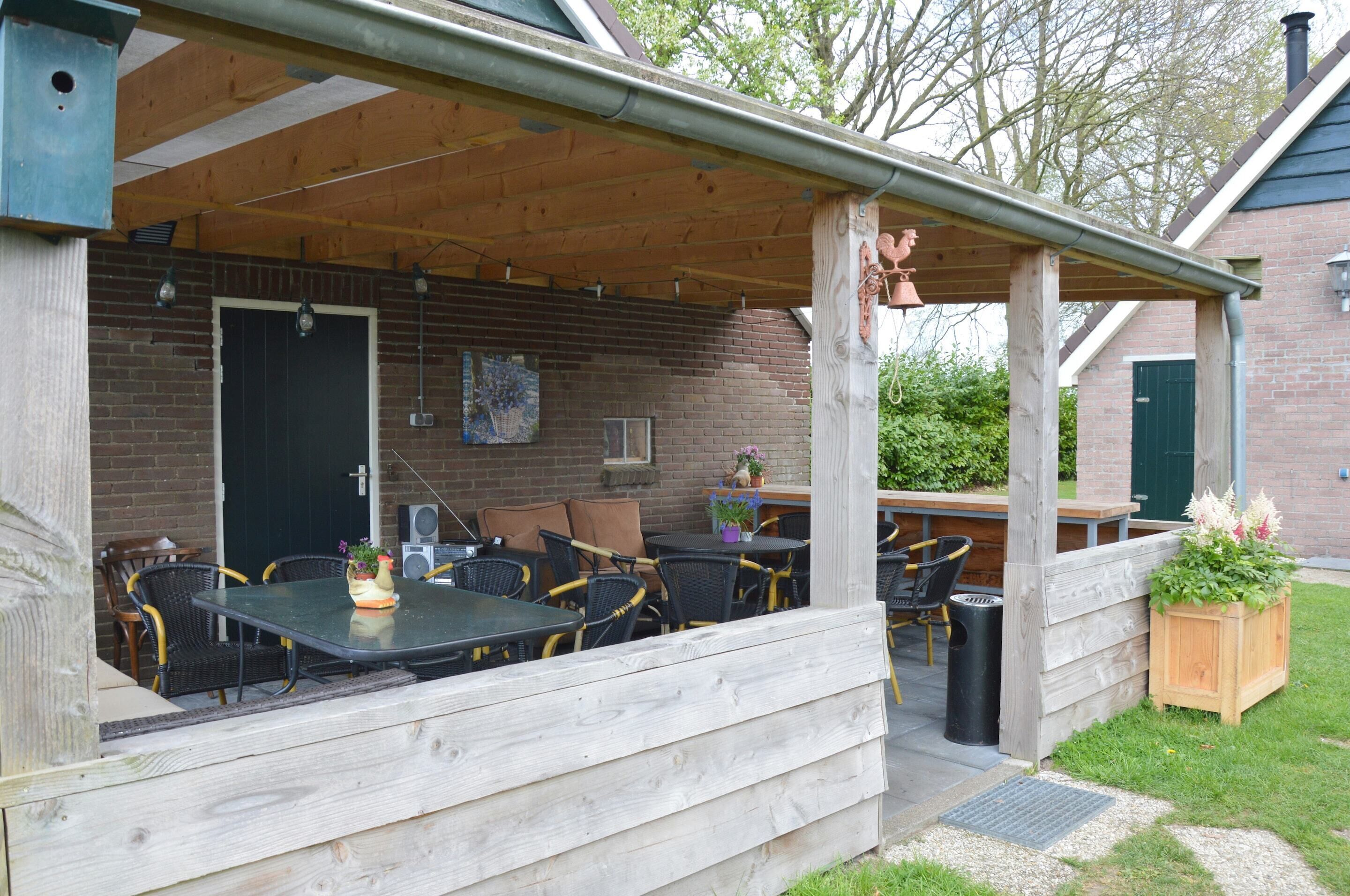 Een terras buiten met tafels en stoelen, een houten hek en een bakstenen gebouw op de achtergrond.
