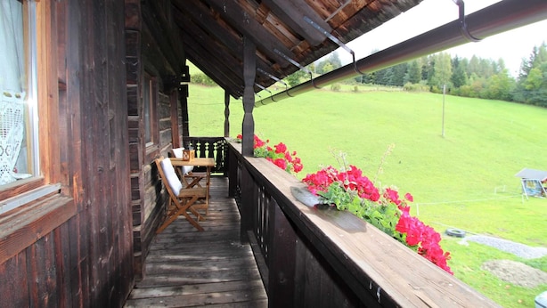 En veranda av trä med ett bord och stolar, ett fönster med en gardin och utsikt över en grön äng med röda blommor.