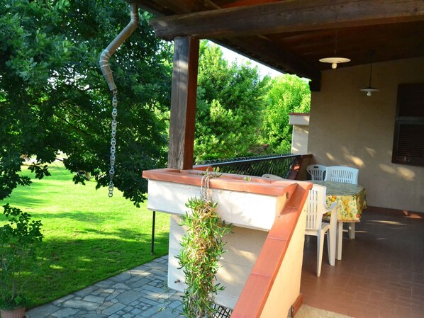 A covered outdoor area with a table and chairs, a hanging light fixture, and a view of a green lawn and trees. - Belvilla by OYO Serena (Massa)