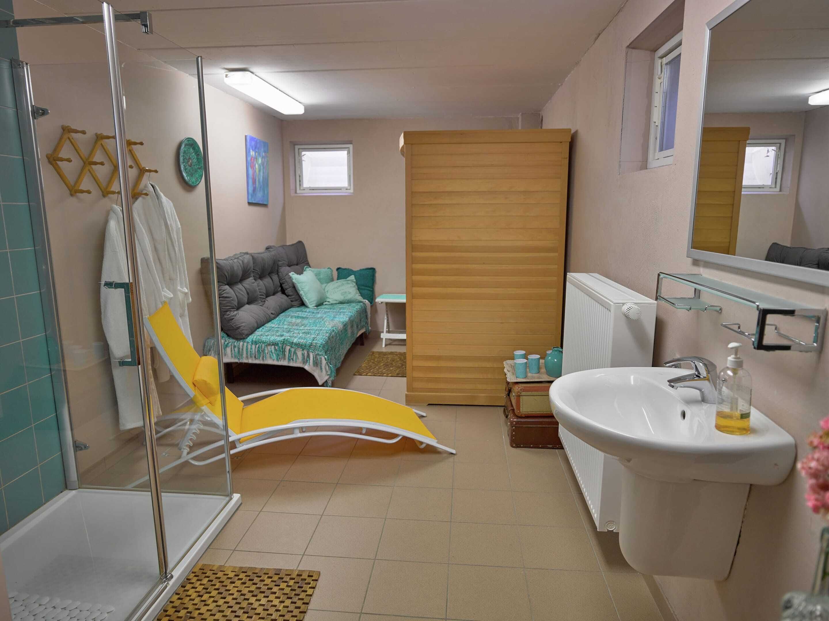 A bathroom with a shower, sink, and toilet. There is a bed with a gray headboard and a yellow chair.