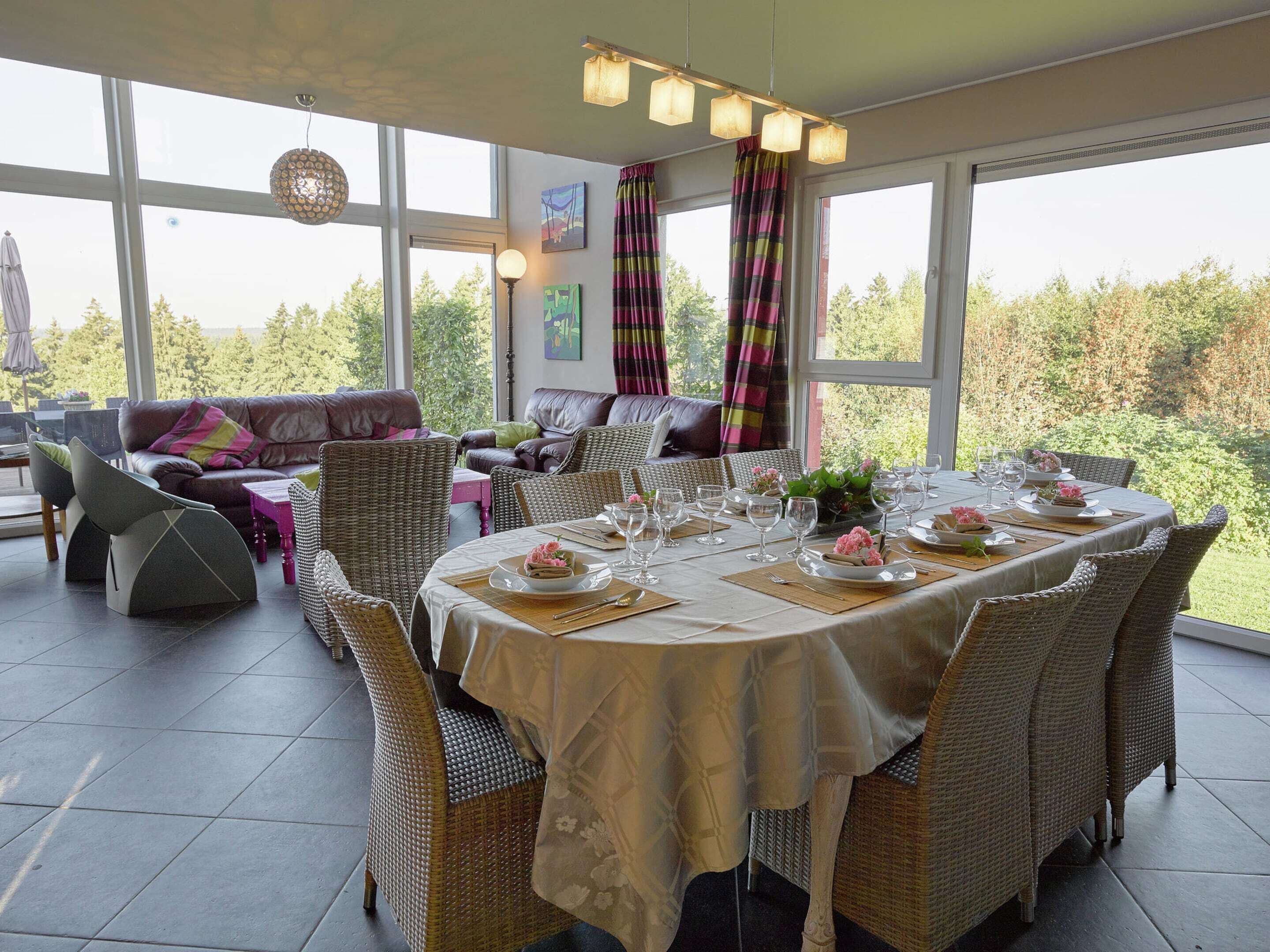 Uma sala de jantar com uma mesa posta para seis pessoas, rodeada por cadeiras de vime, e vista para árvores através de grandes janelas.