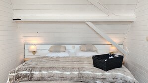 A bedroom with a sloped ceiling, a bed with a fur-like cover, two bedside lamps, and a black basket on the bed. - Pleasant Holiday Home in West Flanders with Enclosed Garden & Hot Tub (Pittem)