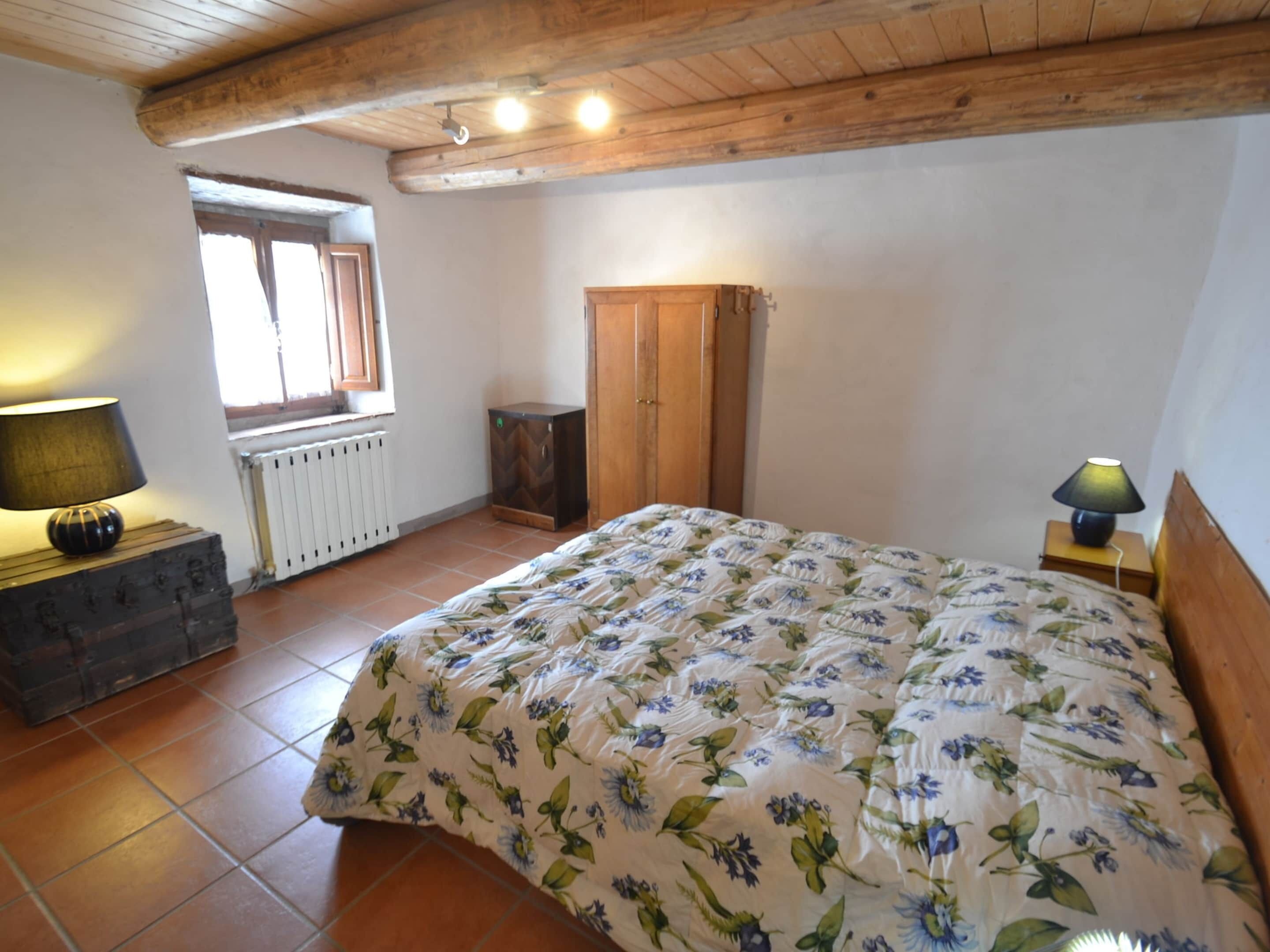 A bedroom with a wooden ceiling, a bed with floral bedding, a radiator, a wooden wardrobe, and a lamp on a nightstand.