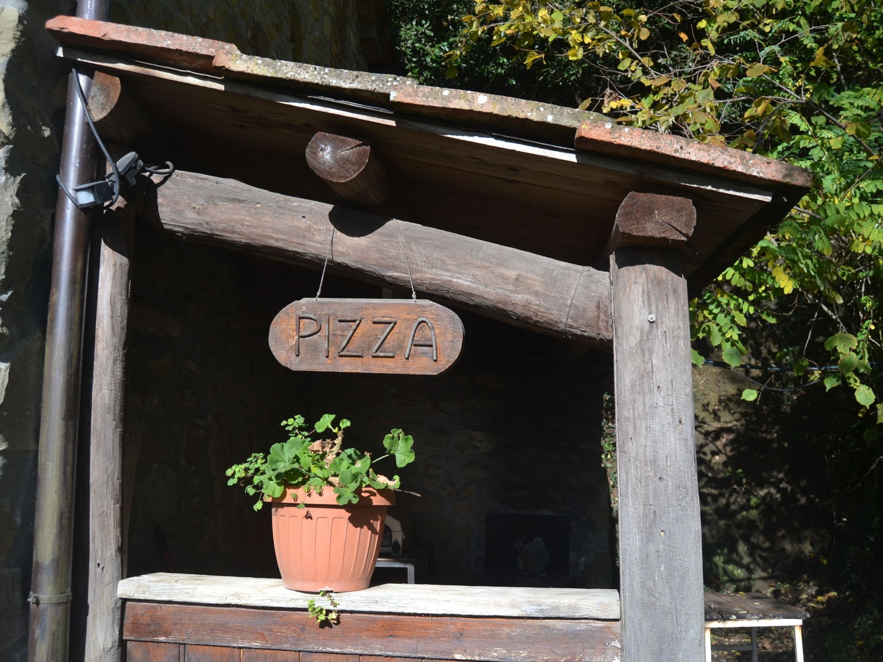 A rustic wooden structure with a 'PIZZA' sign hanging from it, a potted plant on a ledge below, and a stone wall in the background.