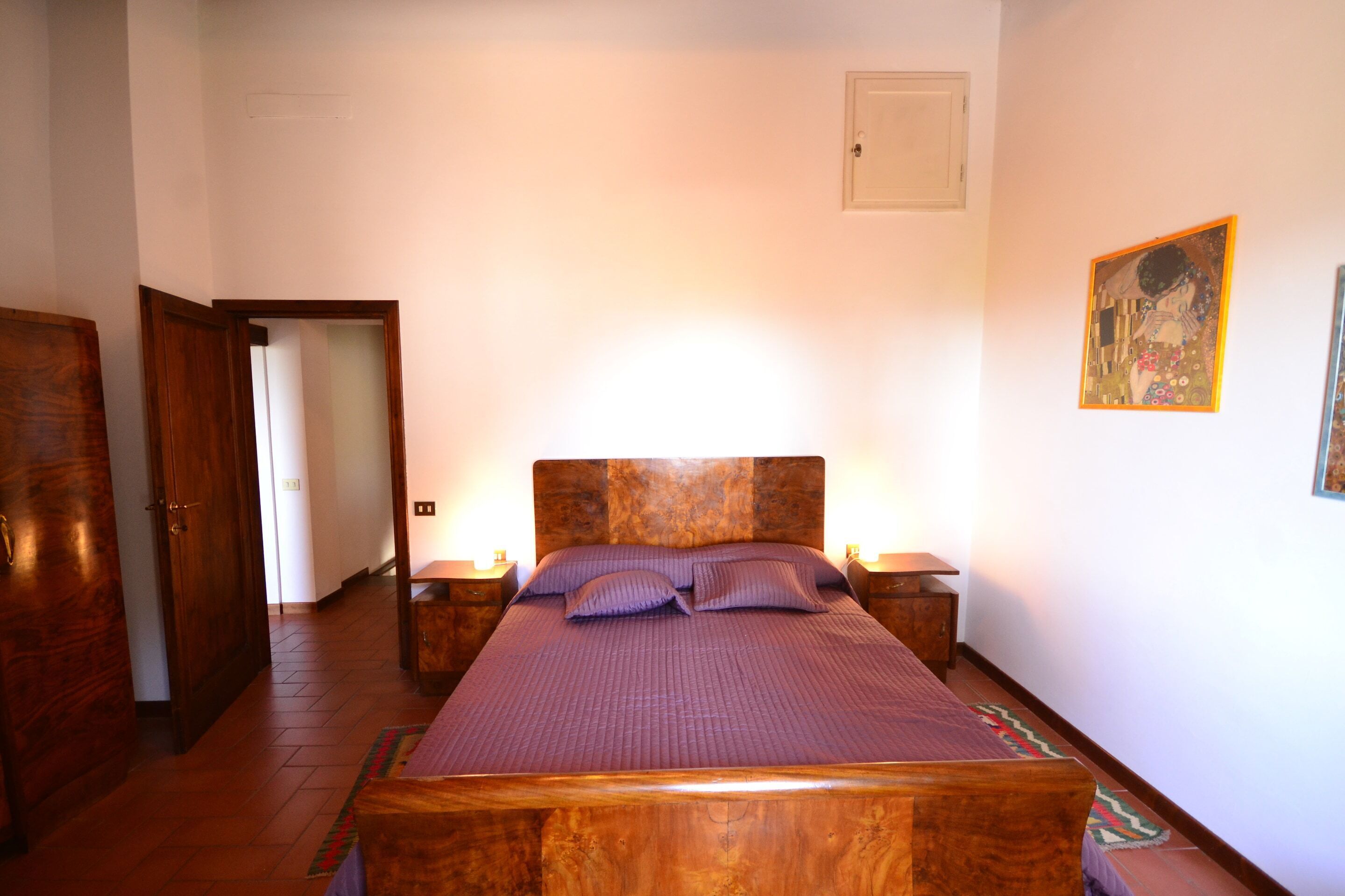 A bedroom with a wooden bed, nightstands, a door, and a wall-mounted artwork.