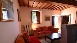 A cozy living room with a sofa, a coffee table, and a television. There are windows with curtains, a radiator, and framed pictures on the walls.