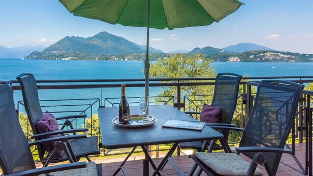 Una terrazza con tavolo, sedie e ombrellone affacciata su uno specchio d'acqua e montagne.