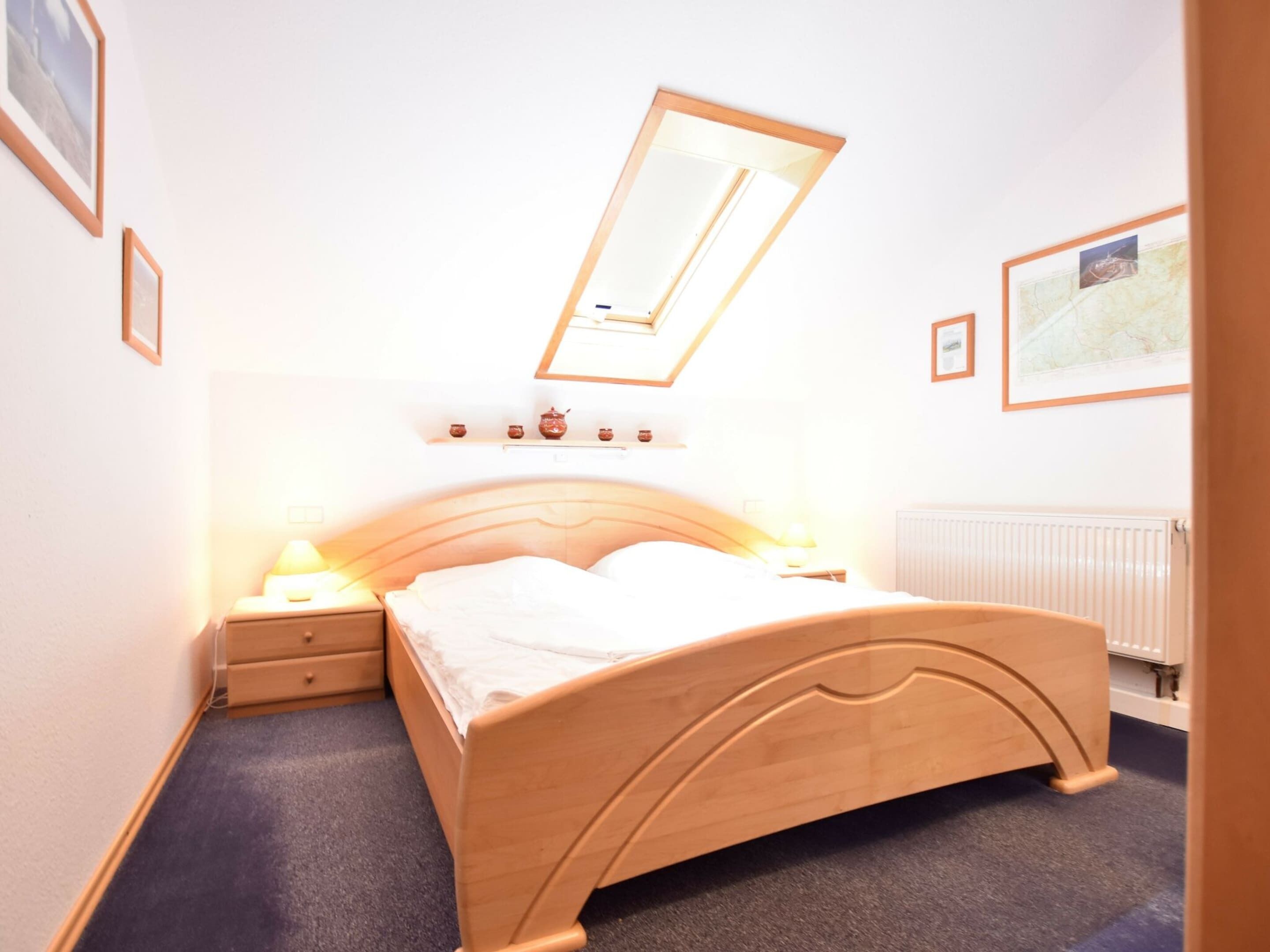 A bedroom with a wooden bed, a nightstand, a bedside table with a lamp, a ceiling light, and a radiator.