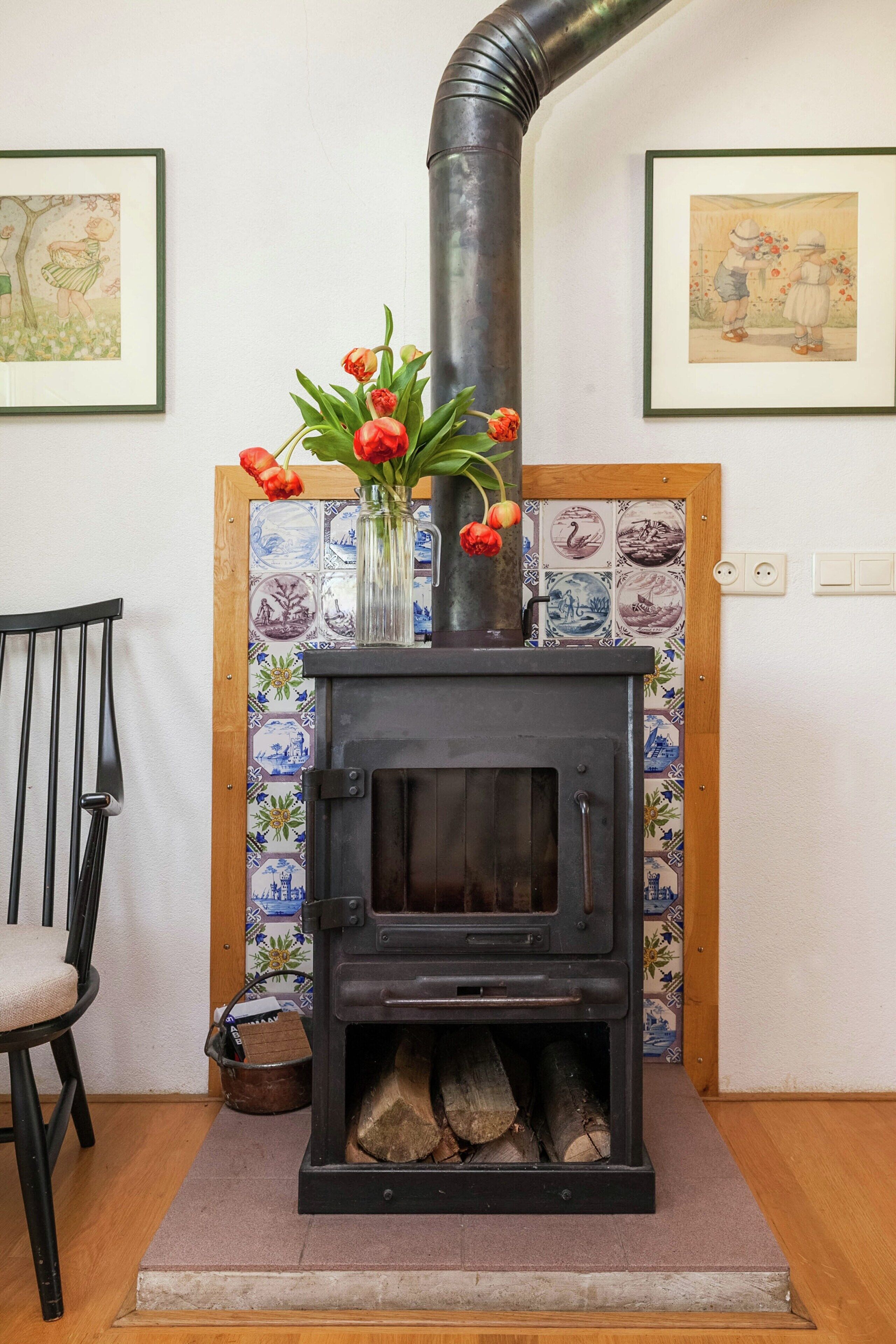 A wood-burning stove with a vase of flowers on top, a chair, and a tiled wall with artwork.