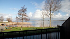 A lakeside view from a balcony with a wooden deck, a grassy area, and a small shed. - Charming Apartment in Langweer With Jetty (Langweer)