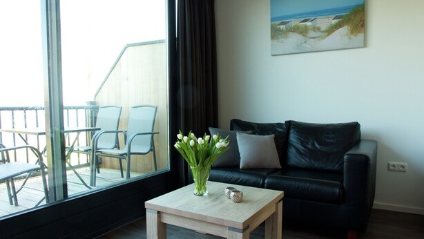 A living room with a glass door leading to an outdoor area, a black leather sofa, a small wooden coffee table with a vase of flowers, and a painting on the wall. - Charming Apartment in Langweer With Jetty (Langweer)