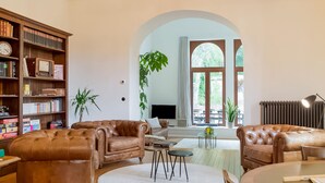 A spacious living room with leather sofas, a bookshelf, a TV, and a large window with a view of the outdoors. - Beautiful Villa With Sauna in Chiny (Chiny)