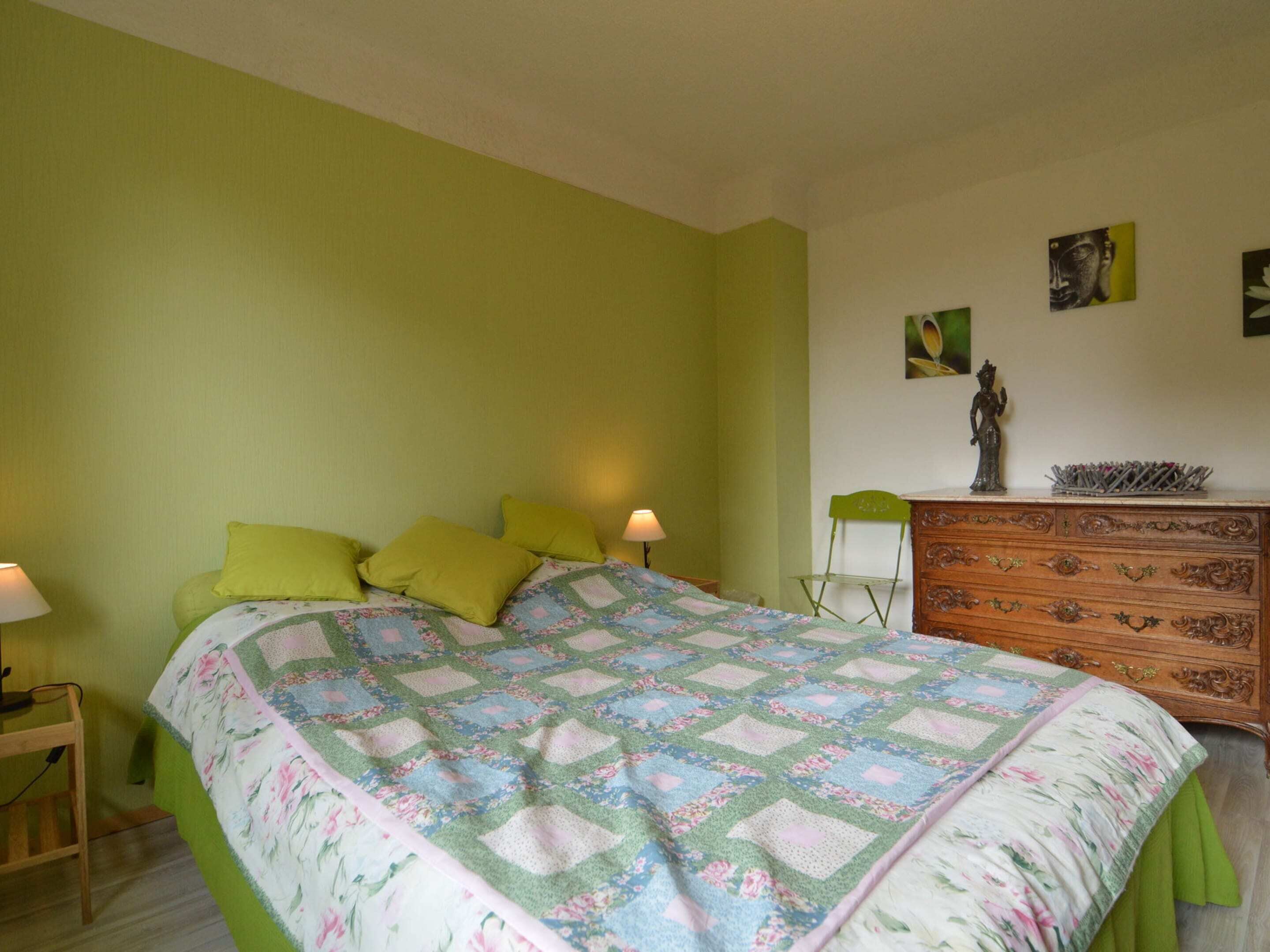 Ein Schlafzimmer mit einem Bett, grünen Wänden, einer Holzkommode und einem Nachttisch mit Lampe.
