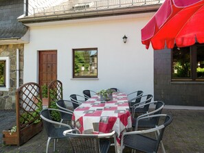 Eine Außenpatio mit einem Tisch, bedeckt mit einer karierten Tischdecke, umgeben von Korbstühlen, einer Holztür und einem roten Sonnendach.