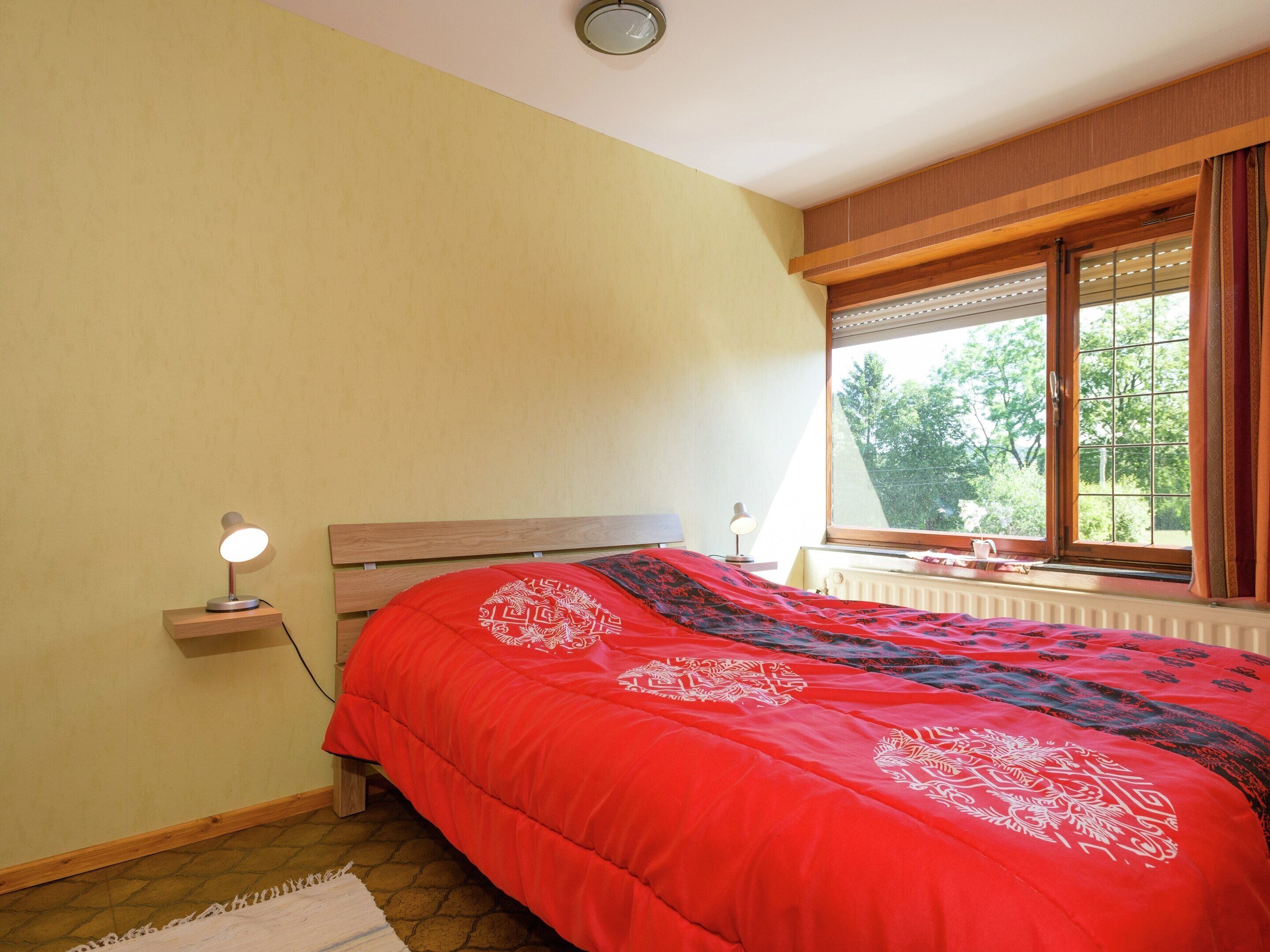 A bedroom with a wooden headboard, a red bedspread with white patterns, a bedside lamp, and a window with curtains.