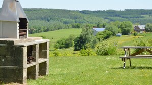 A rustic outdoor setting with a stone barbecue grill, a picnic table, and a view of a rural landscape with houses and trees. - Family Apartment; Bouillon and Semois River (Bievre)