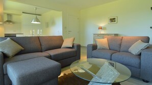 A modern living room with two grey sofas, a glass coffee table, and a kitchen in the background. - Family Apartment; Bouillon and Semois River (Bievre)