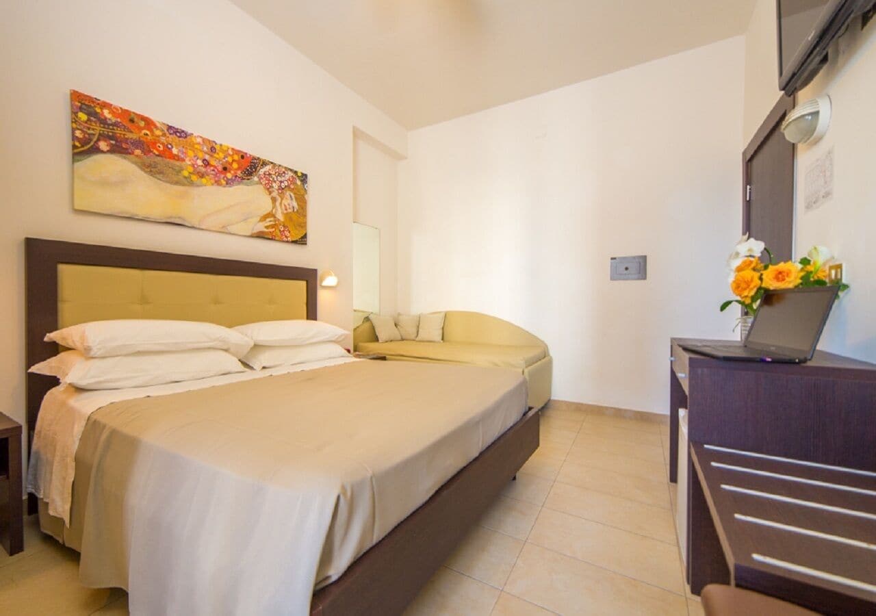 A bedroom with a large bed, a sofa, a desk with a laptop, and a painting on the wall.