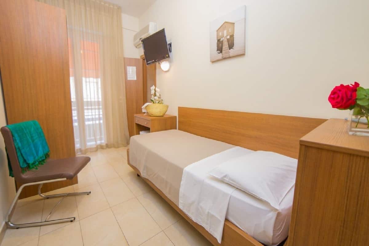 Een hotelkamer met een enkel bed, een houten nachtkastje, een stoel met een groene handdoek, een televisie aan de muur en een foto boven het bed.
