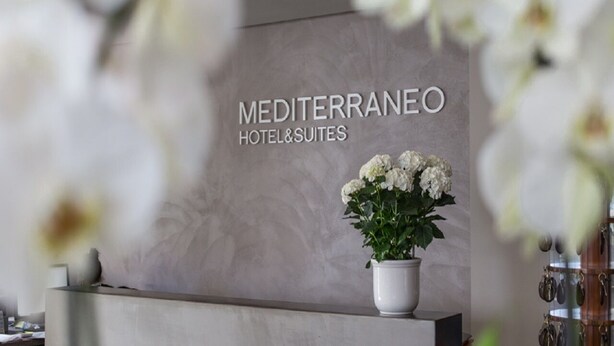 Un comptoir d’accueil avec une enseigne indiquant « Hôtel et appartements Mediterraneo » et une plante en pot ornée de fleurs blanches.