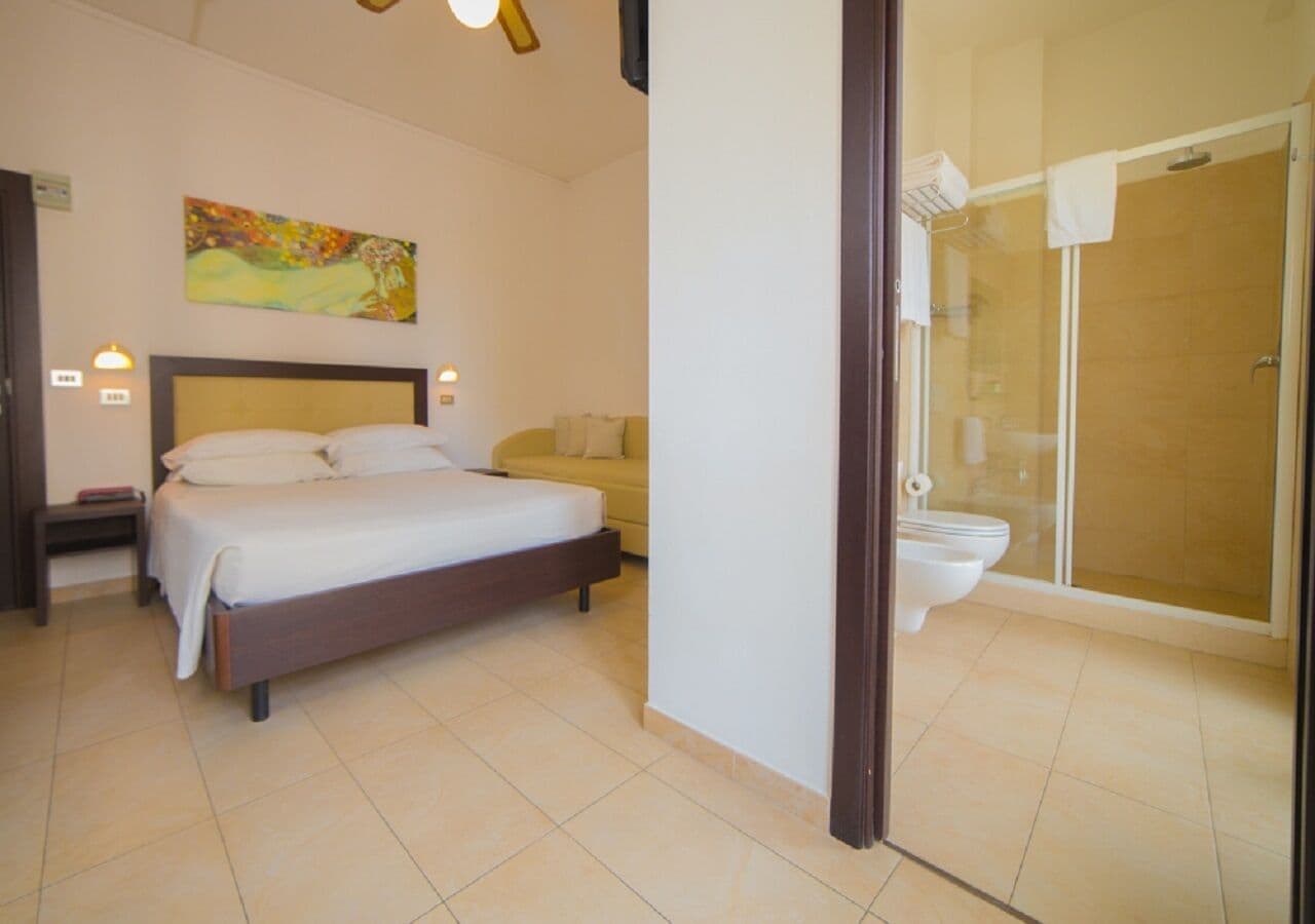 A bedroom with a bed, a bedside table, a nightstand, a sofa, and a shower area with a glass door.