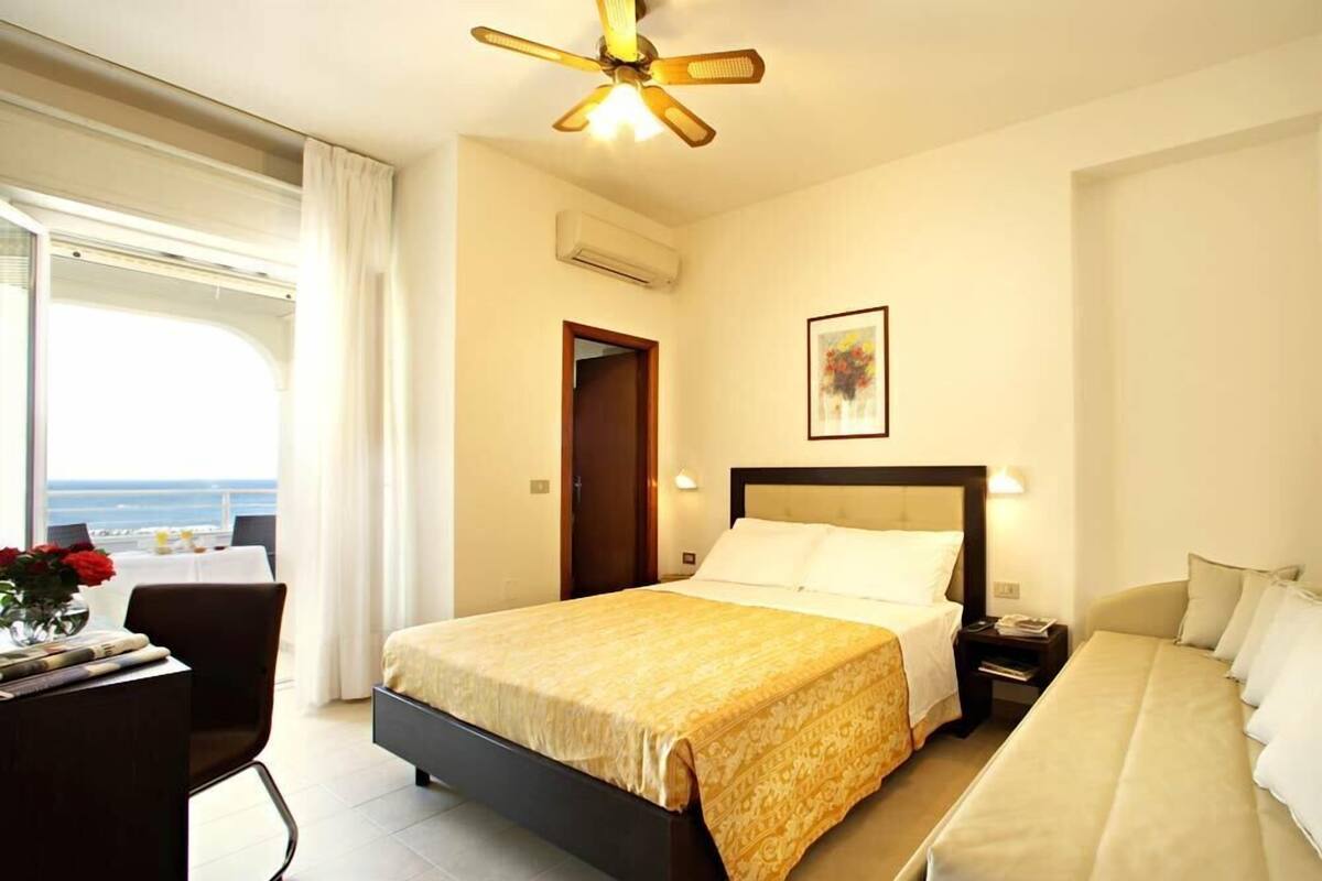 Een hotelkamer met een bed, nachtkastje en uitzicht op zee.