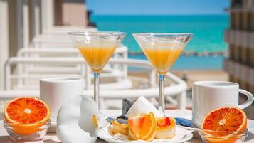 Desayuno con dos vasos de martini de jugo de naranja, un plato con fruta y pasteles, y una taza blanca sobre una mesa junto a una piscina, con un cielo azul claro y el océano al fondo.