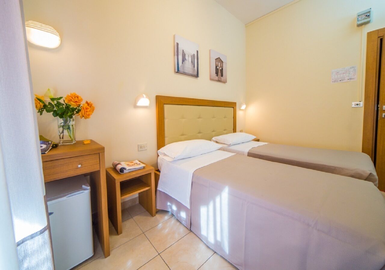 A hotel room with two single beds, a small desk with a chair, a nightstand, a refrigerator, and a vase with flowers.
