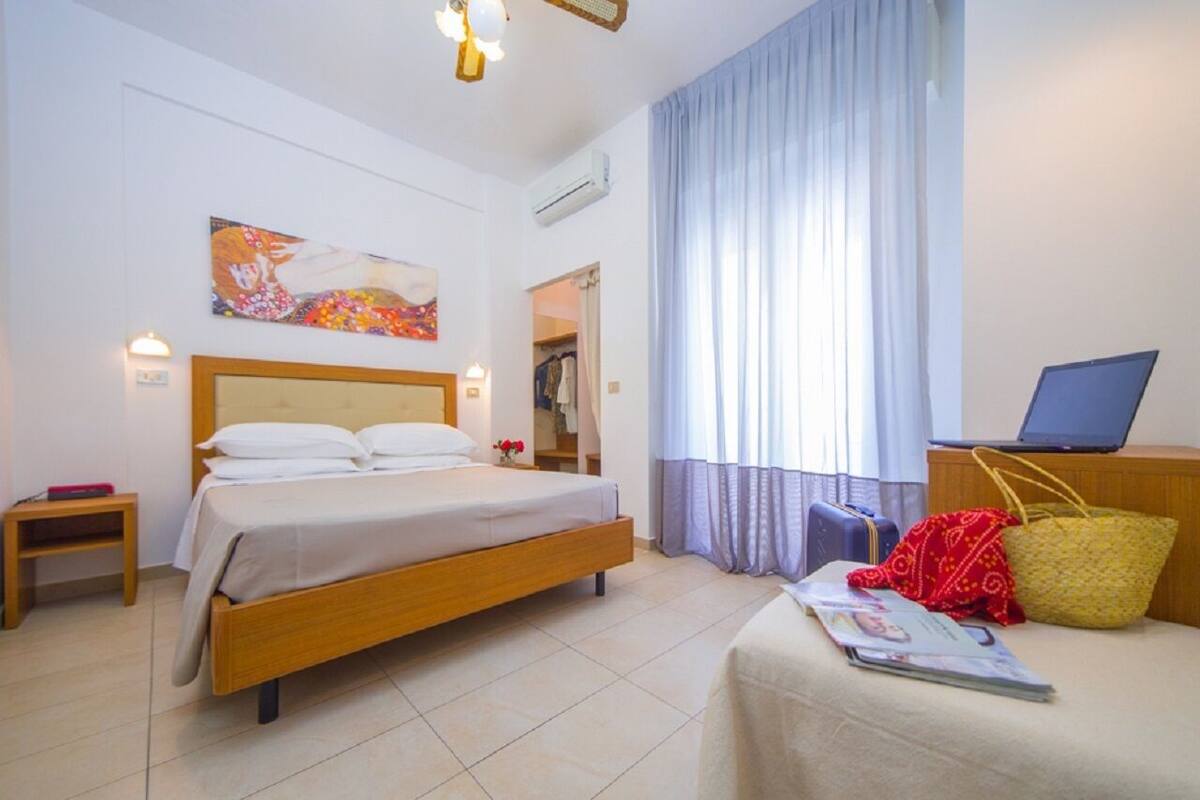 Een hotelkamer met een bed, een bureau met een laptop, een raam met gordijnen en een schilderij aan de muur.