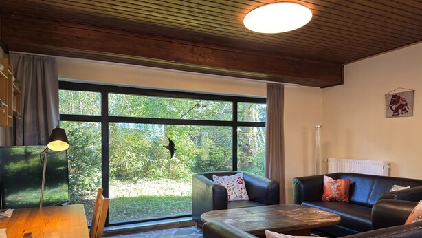 A room with a stone floor, a wooden table, two leather sofas, a lamp, and a large window with a view of greenery. - Grosses Ferienhaus Fuer 8 Personen, Manderscheid (Manderscheid)