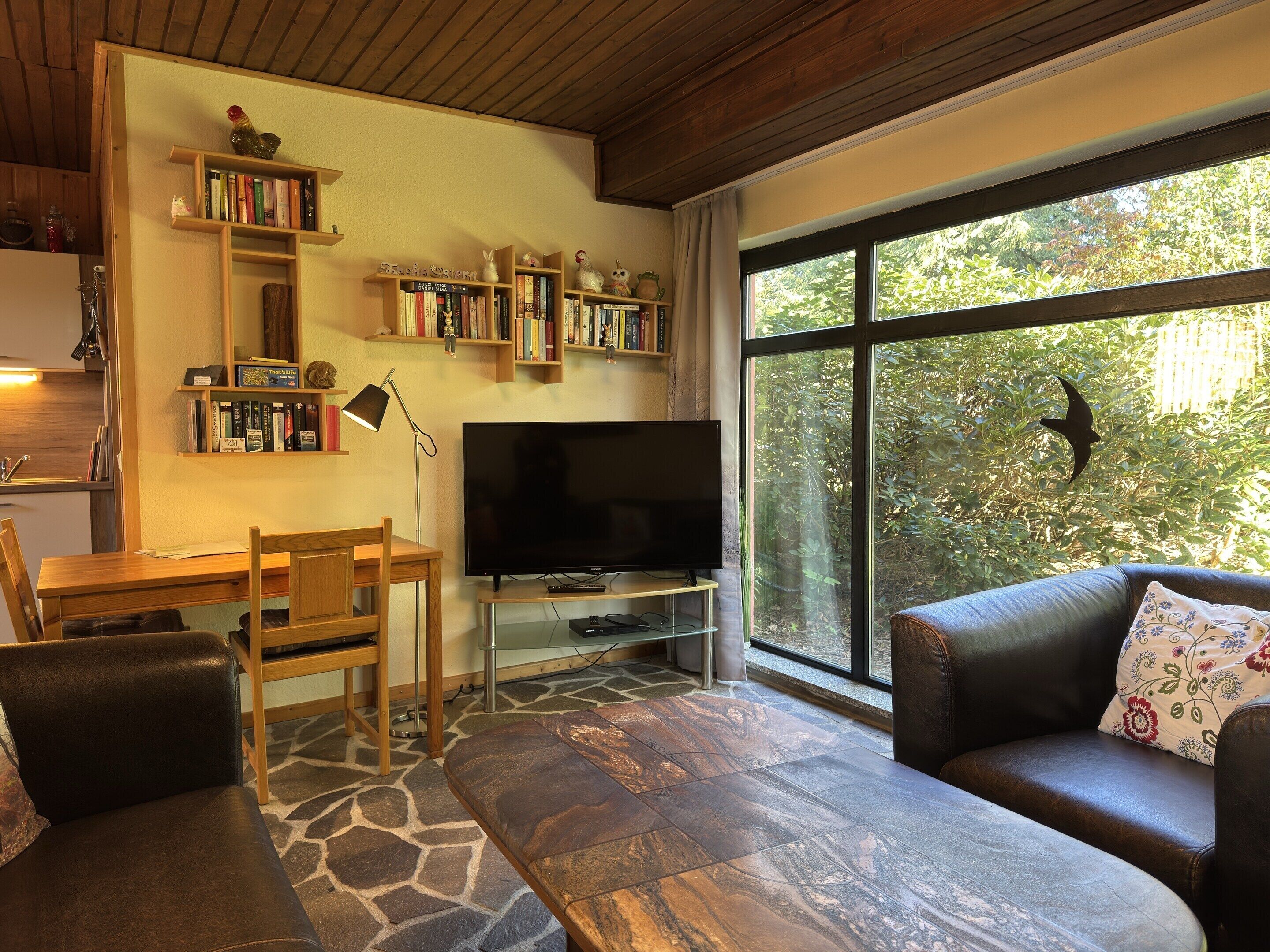 A living room with a stone-patterned floor, a television, a wooden desk, bookshelves, a leather sofa, and a large window with a view of greenery.