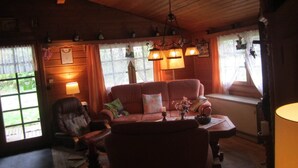 A cozy cabin-style room with wooden walls and ceiling, a leather sofa, a wooden table, and a window with curtains. - Detached Wooden House with Fireplace & Large Garden near Bad Wildungen (Haina)