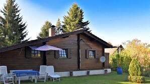 A wooden cabin with a patio area, including a table and chairs, surrounded by a grassy lawn and trees. - Detached Wooden House with Fireplace & Large Garden near Bad Wildungen (Haina)
