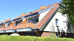 En modern bostadsbyggnad med ett sluttande tak, en balkong med ett räcke och en välskött häck i förgrunden.