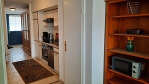 Ett kompakt kök med mikrovågsugn, kaffebryggare och ugn. En hylla i trä används för att förvara köksredskap.
