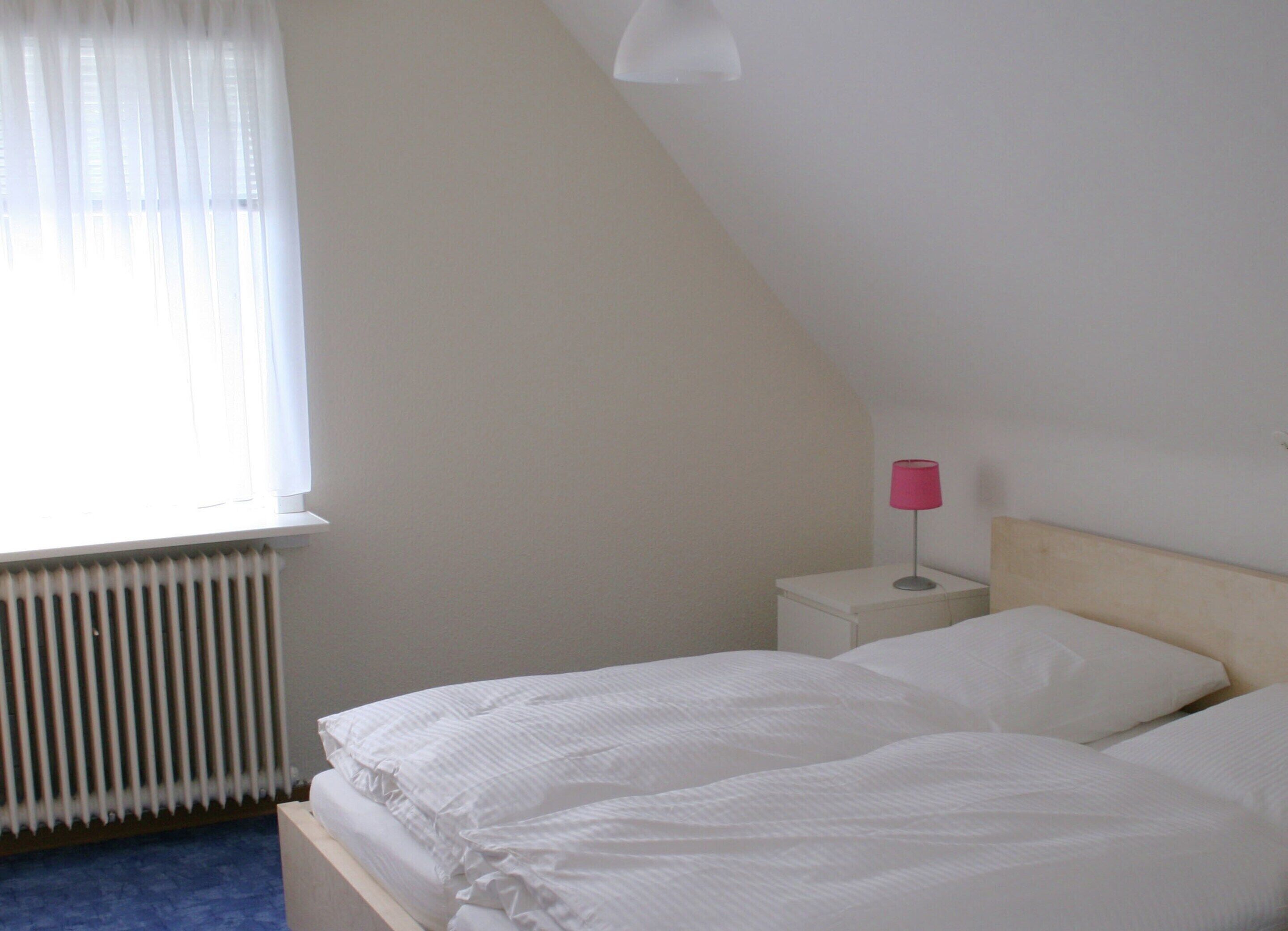 Ein Schlafzimmer mit Bett, Nachttisch, Lampe, Fenster mit Vorhängen und Heizkörper.
