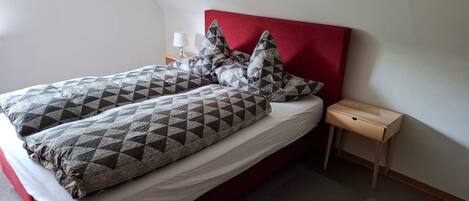 A bed with a red headboard, grey and black patterned bedding, and a small wooden nightstand with a lamp.