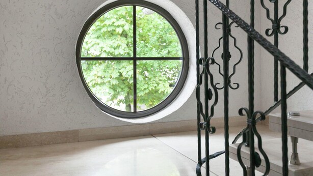 A round window with a four-pane design, a metal staircase with ornate balusters, and a view of green foliage outside. - Eschfeld Sauna Apartment (Eschfeld)