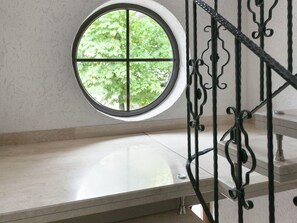 A round window with a four-pane design, a metal staircase with ornate balusters, and a view of green foliage outside. - Eschfeld Sauna Apartment (Eschfeld)