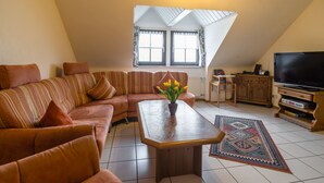 A cozy living room with a sectional sofa, a coffee table, a flat-screen TV, and a large window with curtains. - Eschfeld Sauna Apartment (Eschfeld)