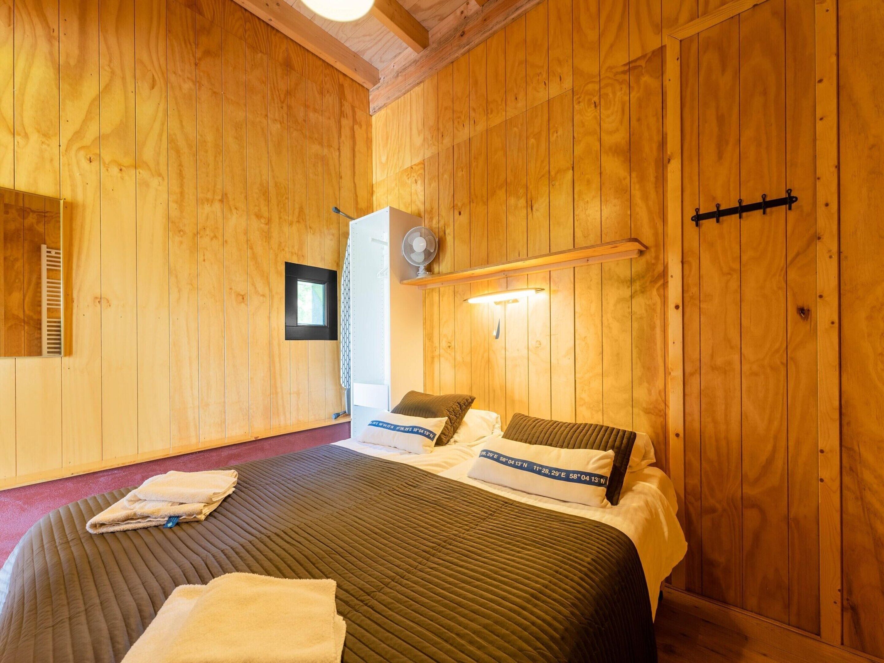 A wooden-paneled room with a bed, a small window, a fan, and a door with a black handle.