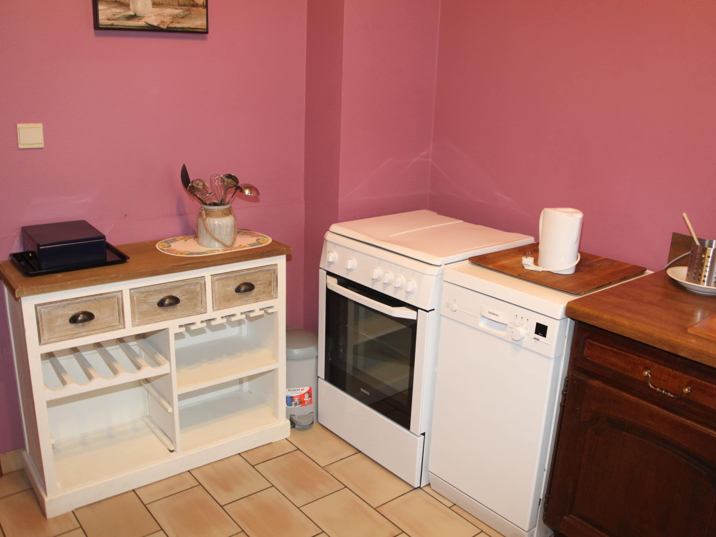 Een kleine keuken met een wit fornuis, oven en vaatwasser, een houten tafeltje met een vaas en een roze muur.